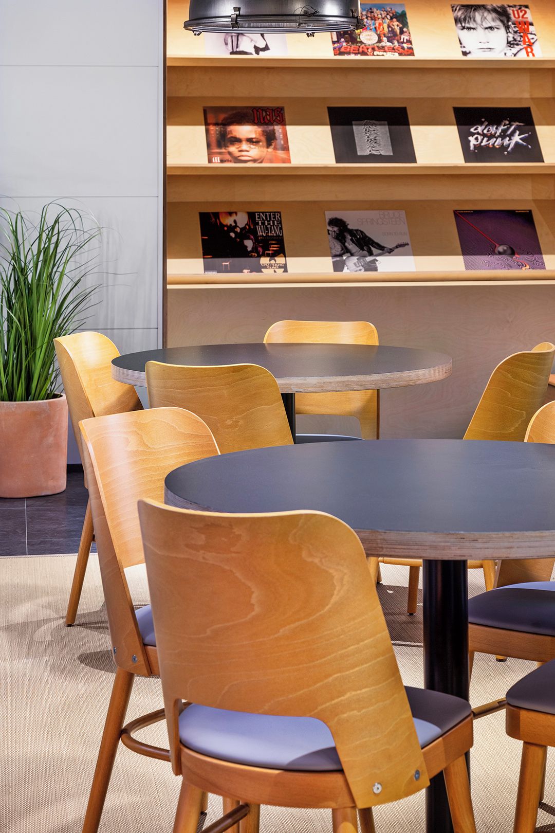 A dining area with round tables and wooden chairs, a potted plant, and a wall with framed posters.