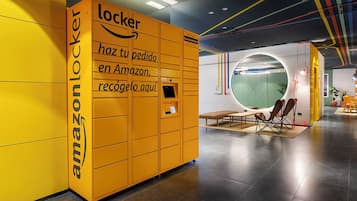 Een gele Amazon Locker met een digitaal scherm, een bank en een rond raam in een modern interieur.