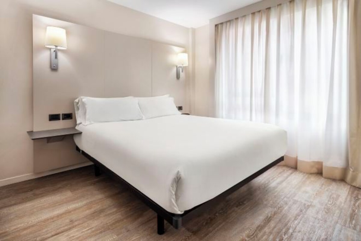 Habitación de hotel con una cama grande, dos lámparas fijadas a la pared, suelo de madera y una ventana con cortinas.