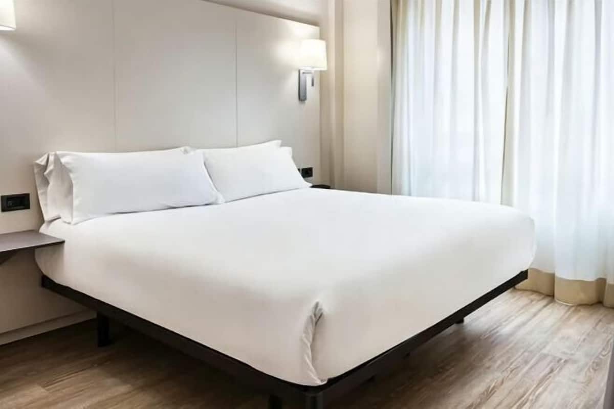Una cama bien hecha con sábanas blancas, una mesita de noche con una lámpara y una ventana con cortinas traslúcidas.