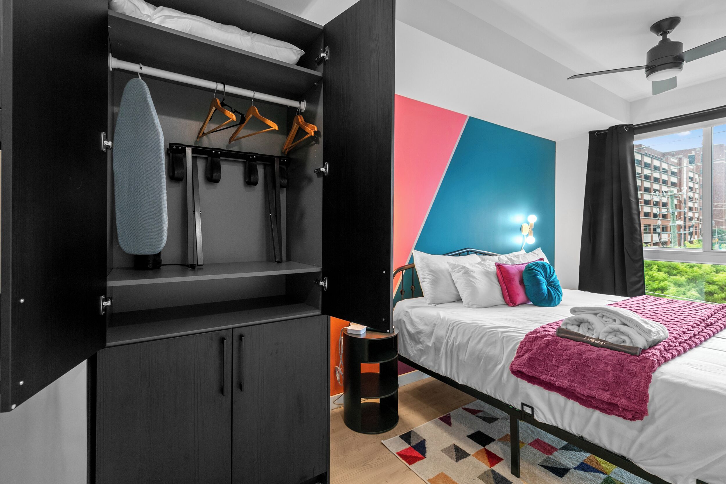 A modern hotel room with a bed, a wardrobe, and a view of the city.