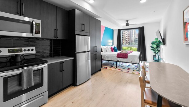 A modern kitchen with stainless steel appliances, a dining area with a wooden table and chairs, and a view of a cityscape through a large window. - King Bed, Gym, 2min walk to UAB, Foodie Haven (Birmingham)