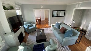 Ein Wohnzimmer mit einer Couch, Sesseln, einem Couchtisch und einem Fernseher. Im Hintergrund ist eine Essecke mit einem Tisch und Stühlen zu sehen.