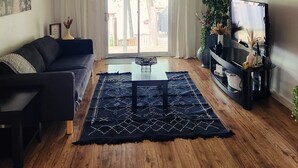 Un salon avec un canapé, une table basse, un téléviseur posé sur un meuble et une grande fenêtre avec des rideaux.