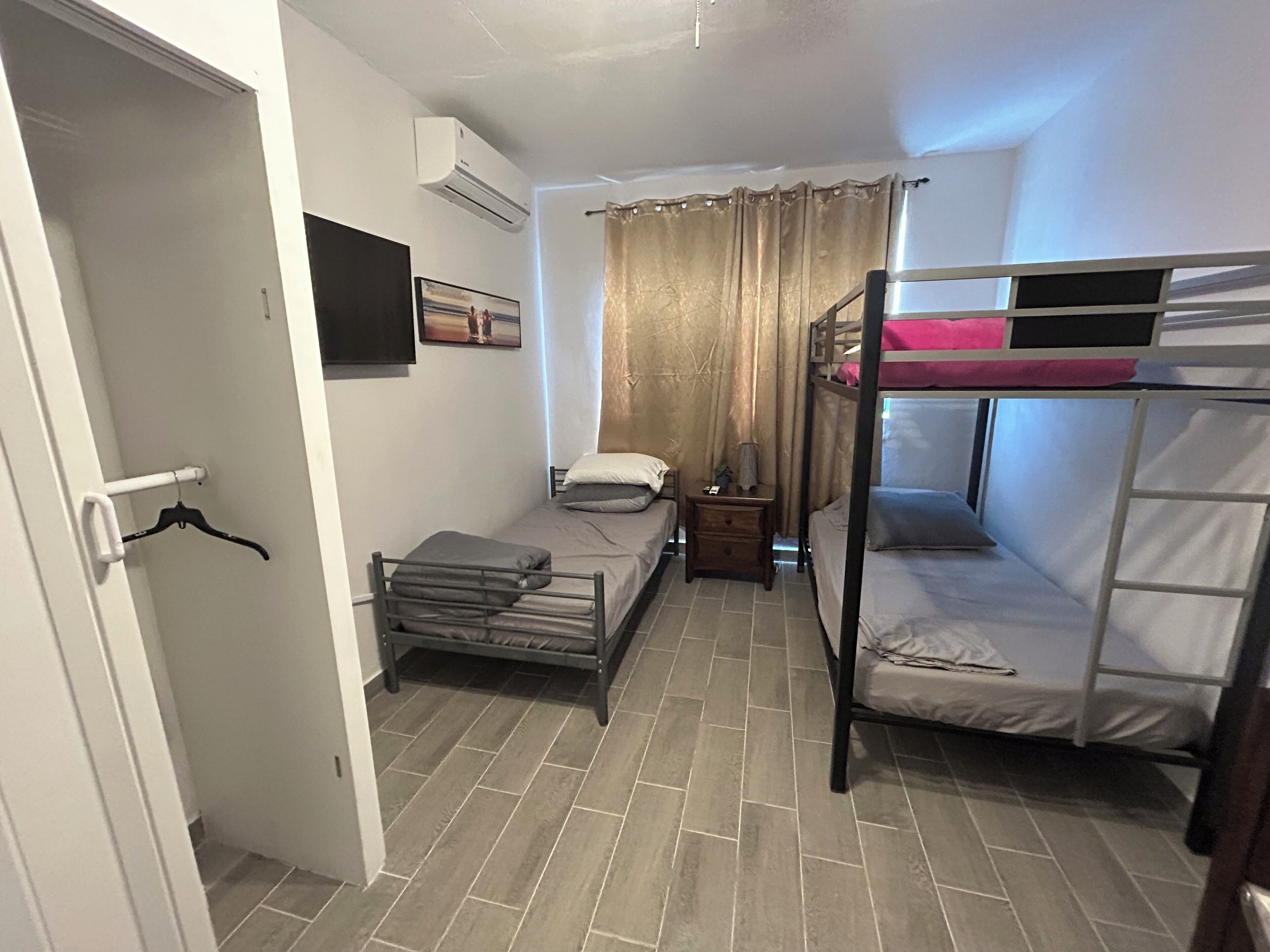 A compact room with bunk beds, a small bedside table, a wall-mounted air conditioner, and a door with a coat rack.