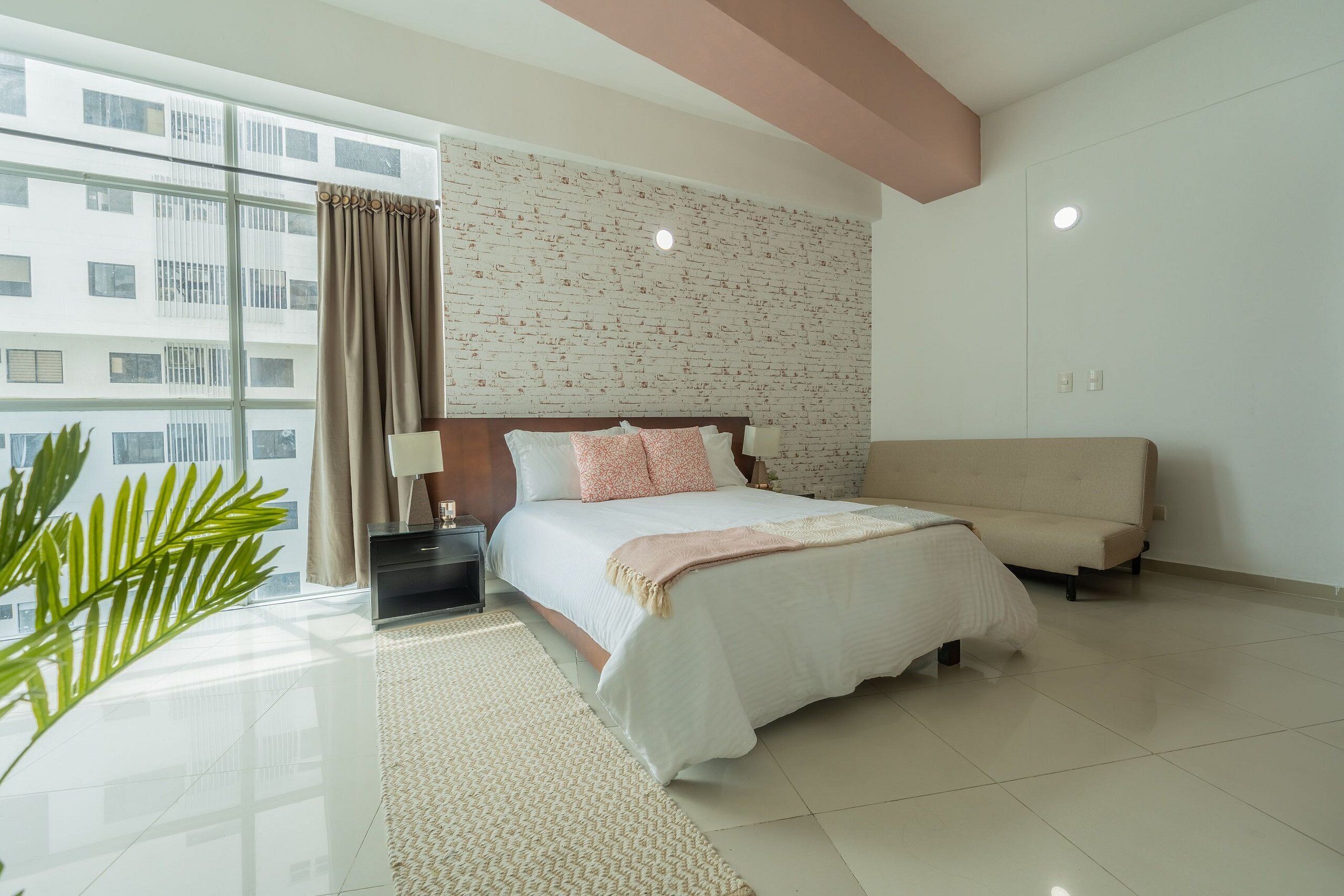 Une chambre d’hôtel moderne, dotée d’un grand lit, d’un canapé, d’une table de chevet, d’une lampe et offrant une vue sur la ville grâce à de grandes fenêtres.