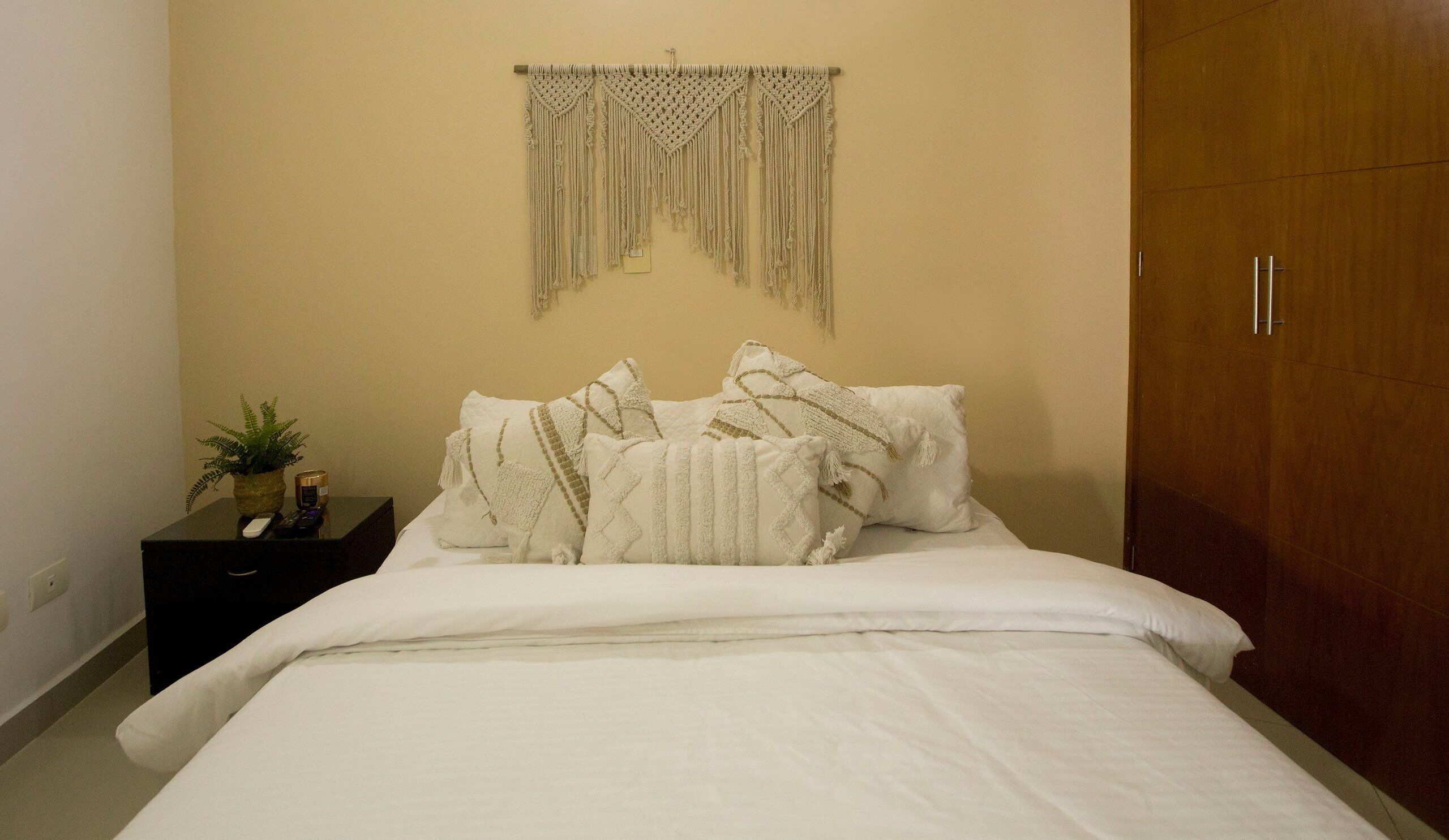 Una cama bien hecha con sábanas blancas y cojines decorativos, una mesita de noche con una planta y una vela, y un colgador de macramé sobre la cama.