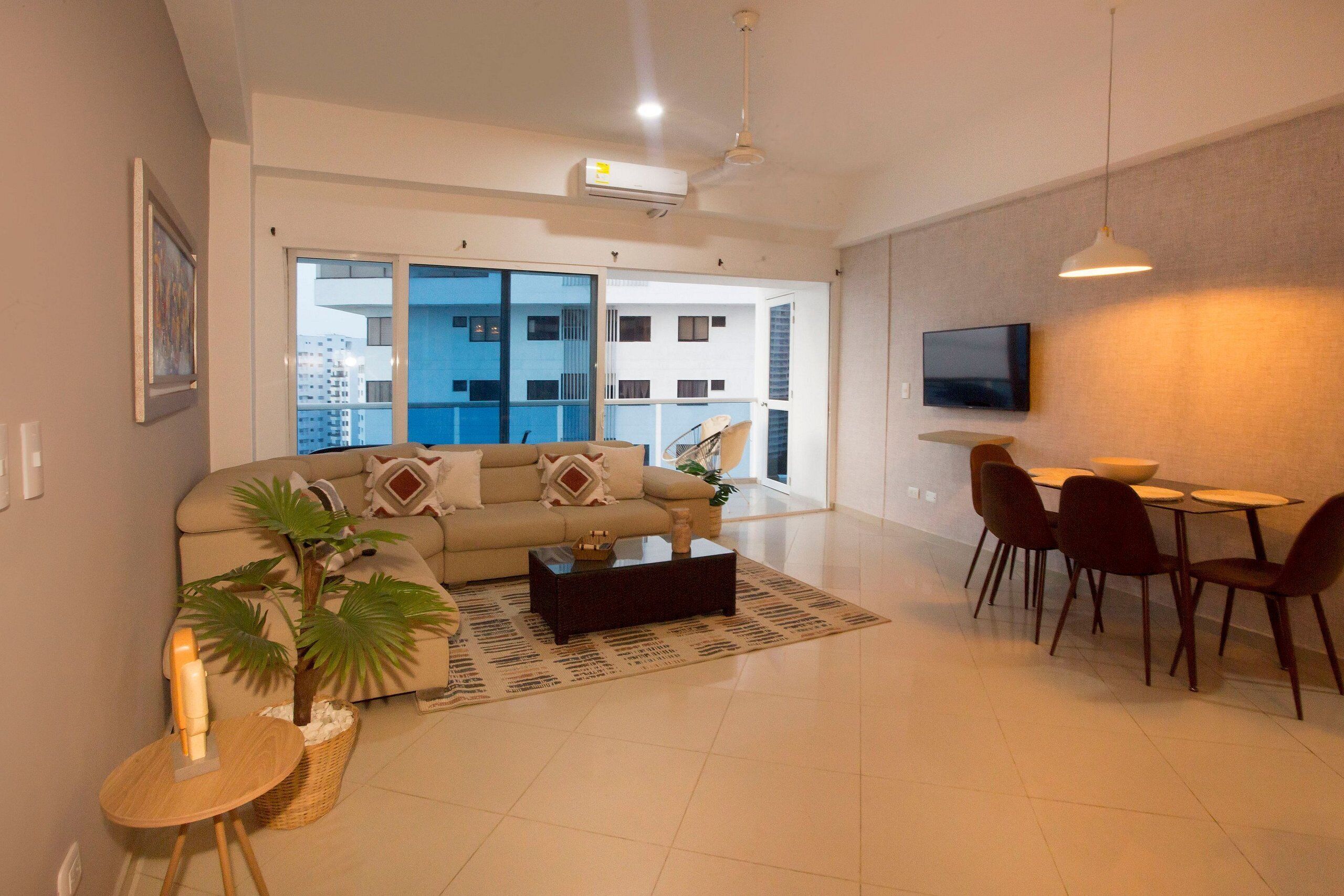 A modern living room with a sofa, coffee table, dining table, chairs, a TV, and a balcony with a city view.