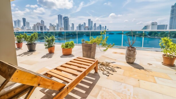 A balcony with a wooden lounge chair, potted plants, and a view of a city skyline and water body. - Poseidon del Caribe (Cartagena)