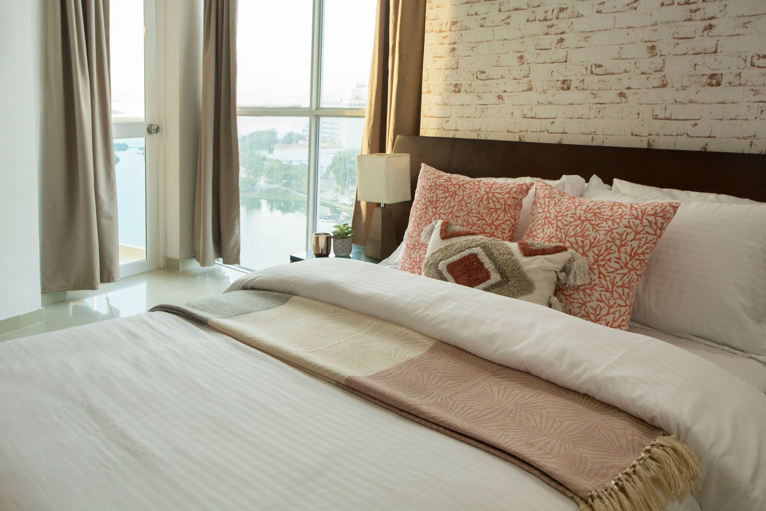 Una habitación de hotel con una cama grande, vista a un cuerpo de agua a través de una ventana y una pared con diseño de ladrillo.
