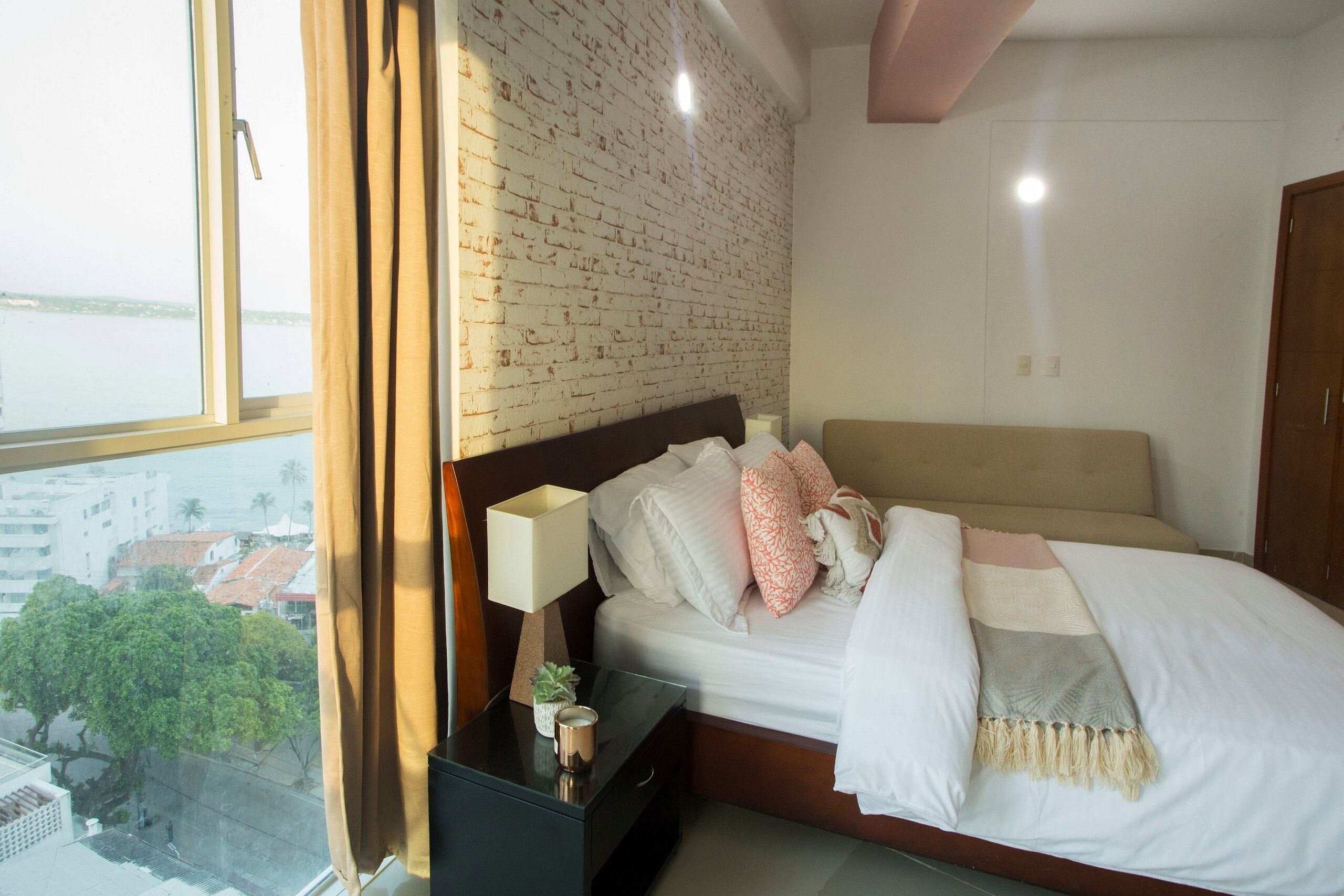 Un dormitorio con pared de ladrillo, cama con ropa de cama blanca, mesita de noche con lámpara y vistas a un paisaje urbano a través de una ventana.