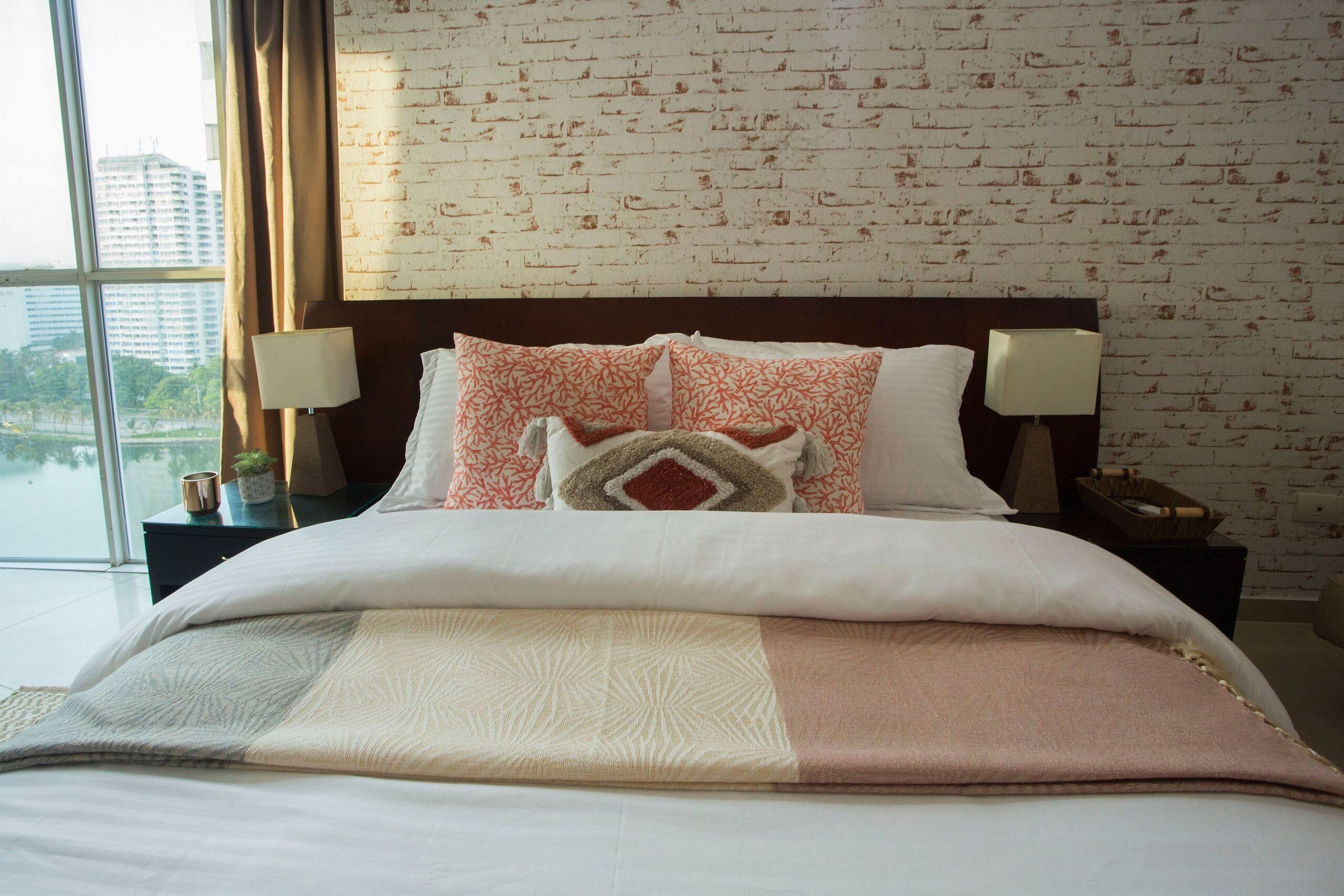 Habitación de hotel con pared de ladrillos, una cama grande con ropa de cama estampada, dos lámparas de noche y vistas a un paisaje urbano a través de un ventanal amplio.