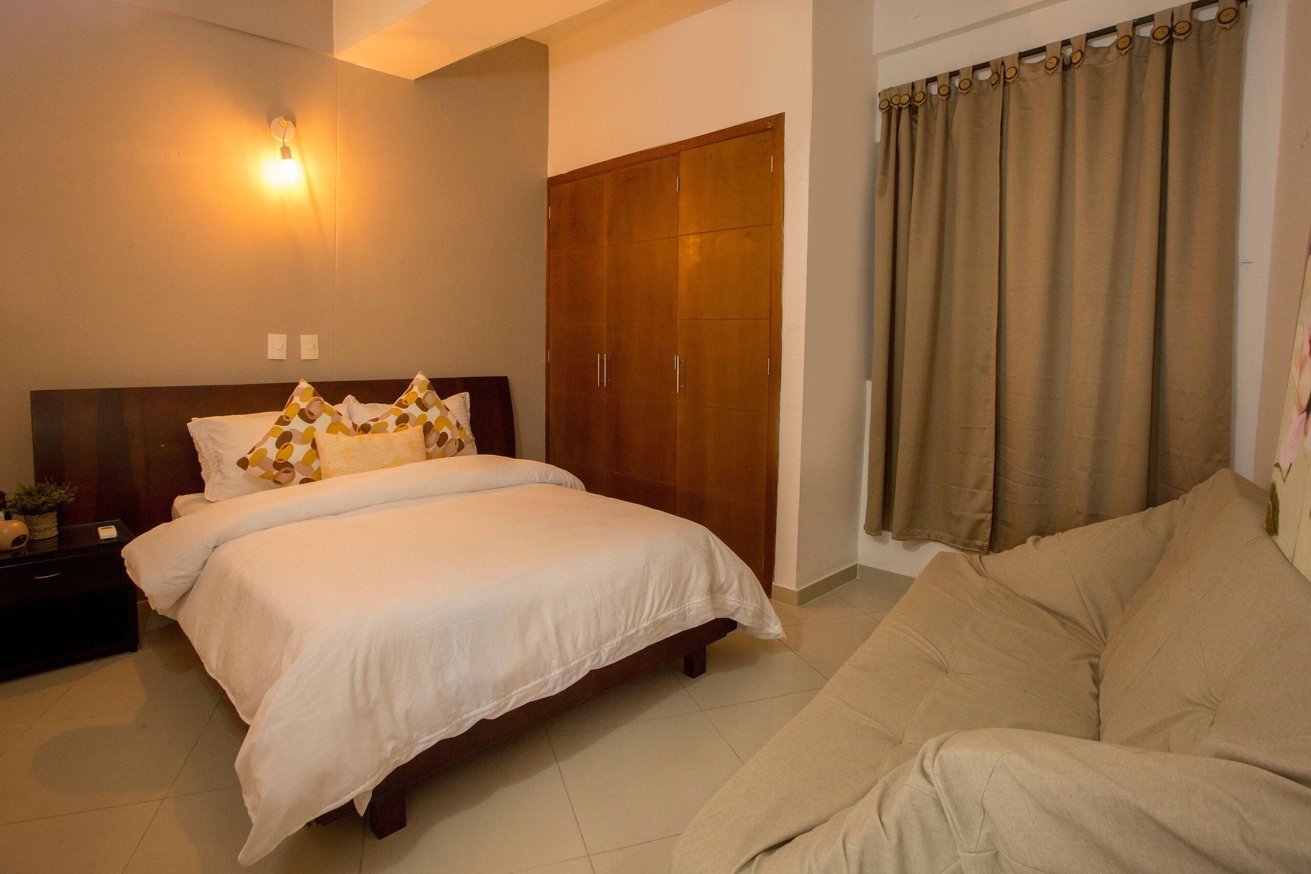 A hotel room with a bed, a wardrobe, a sofa, and a wall-mounted light fixture.