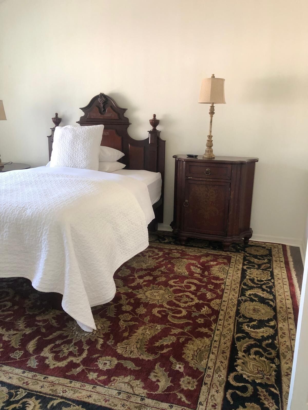 A neatly made bed with white linens, a wooden headboard, and a matching nightstand with a lamp. The room features a patterned carpet and a simple, clean design.