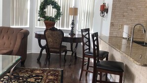 Ein Wohnzimmer mit braunem Sofa, einem Glastisch, einem gemusterten Teppich, einem Essbereich mit Holztisch und Stühlen, einer Küche mit Ziegelrückwand und einem großen Fenster mit Jalousien.