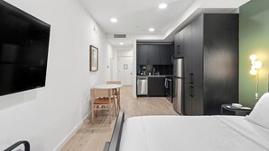Een moderne hotelkamer met een bed, een bureau, een stoel, een klein tafeltje, een televisie, een kitchenette en een groene muur met een lamp.