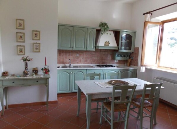 Cuina amb armaris verds, respatller de rajola, finestra amb cortina i taula de menjador amb cadires.