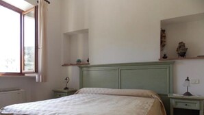 Un dormitorio con cama, mesita de noche, cómoda, ventana y estante con objetos decorativos.