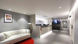 Zona de recepció d'hotel amb un sofà blanc, una catifes vermella, un taulell de recepció, una pantalla de televisió i un quadre amb marc a la paret.