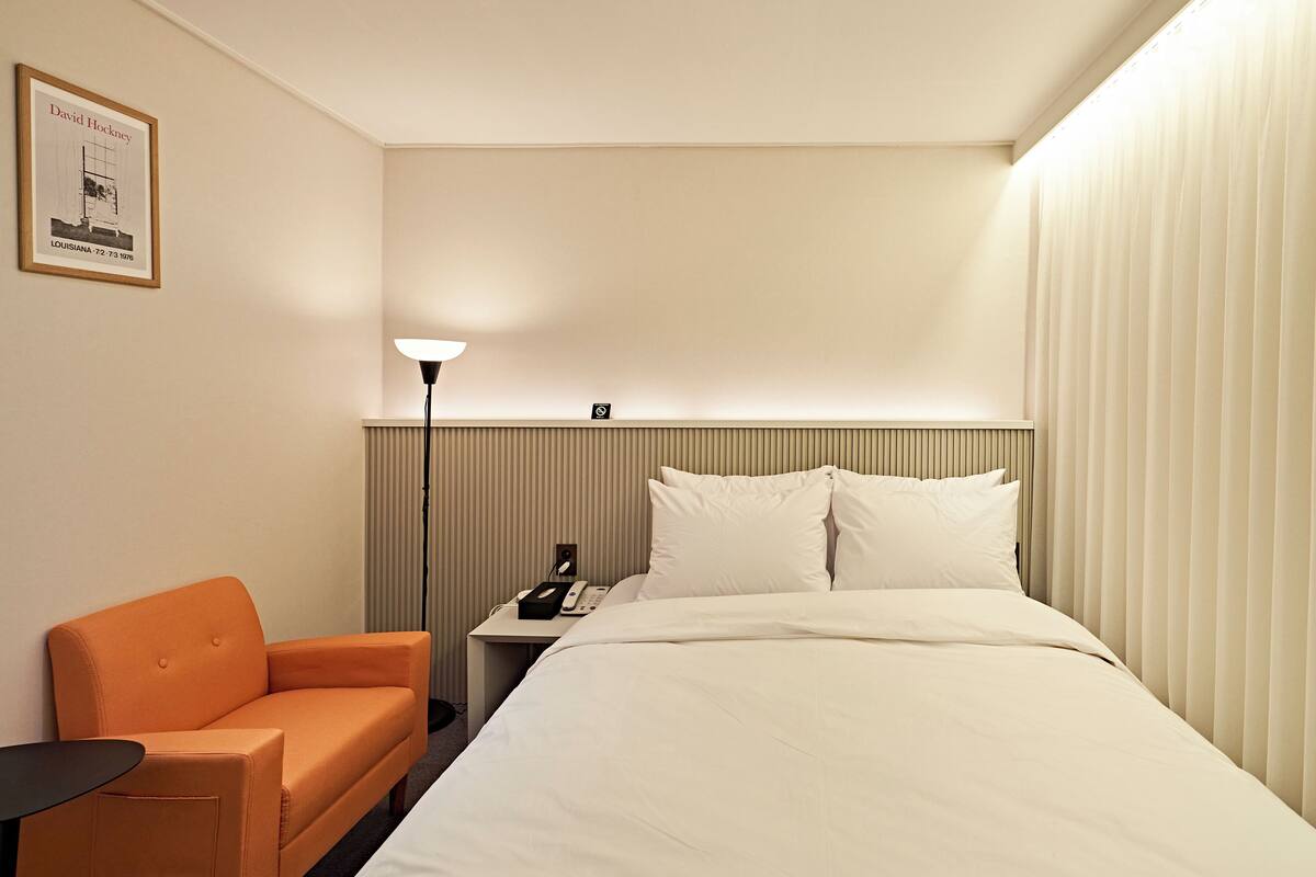 Habitación de hotel con cama, una silla naranja, mesita de noche con lámpara y un cuadro en la pared.