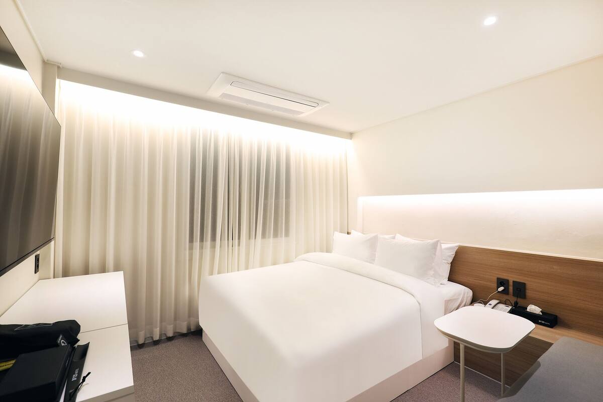 Habitación de hotel moderna con una cama grande, una mesita de noche, un televisor y una ventana con cortinas translúcidas.