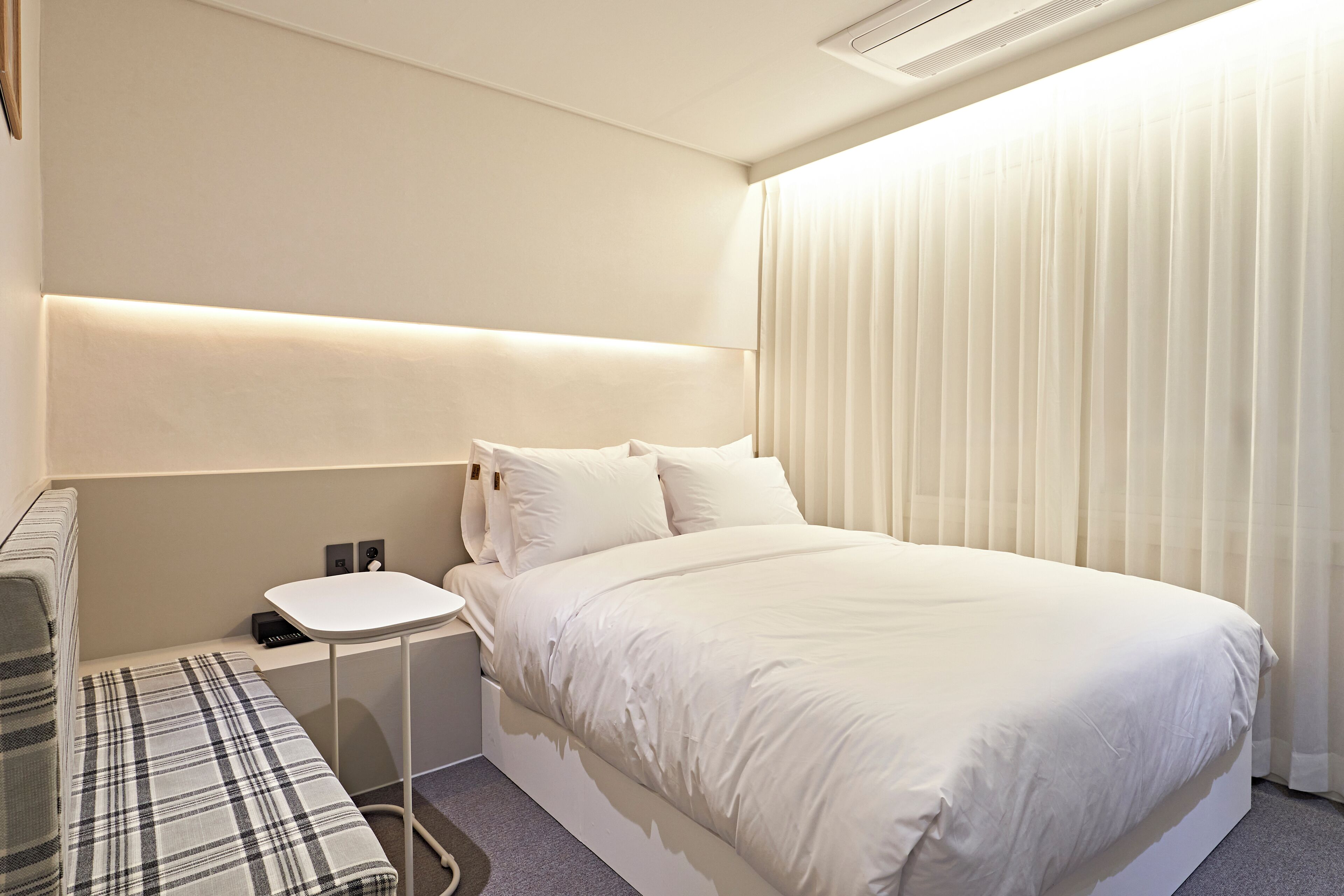 Moderni hotellihuone, jossa on siististi pedattu sänky, yöpöytä ja penkki.