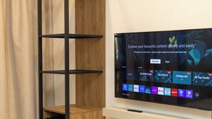 Un salon moderne avec un téléviseur posé sur un meuble blanc, une étagère en bois et un rideau.
