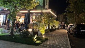 Un bâtiment moderne doté d’un espace extérieur bien éclairé, comprenant un chemin pavé et un espace de détente avec des tables et des chaises.