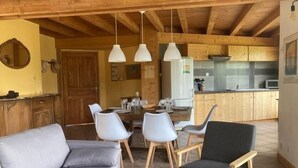 Ein gemütlicher Wohnbereich mit einer Couch, Sesseln und einem Esstisch für vier Personen. Im Hintergrund ist eine Küche mit Holzschränken und einem Kühlschrank zu sehen.