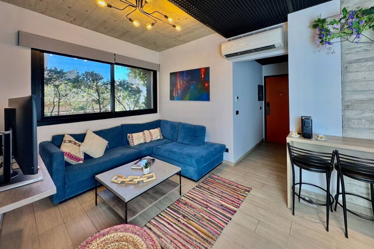 Una sala de estar moderna con un sofá azul, una mesa de centro y un televisor. Se ve una ventana con vistas a árboles, un cuadro en la pared y una zona de comedor con sillas.
