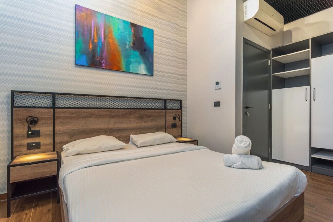 A modern hotel room with a large bed, a wardrobe, and a colorful abstract painting on the wall.
