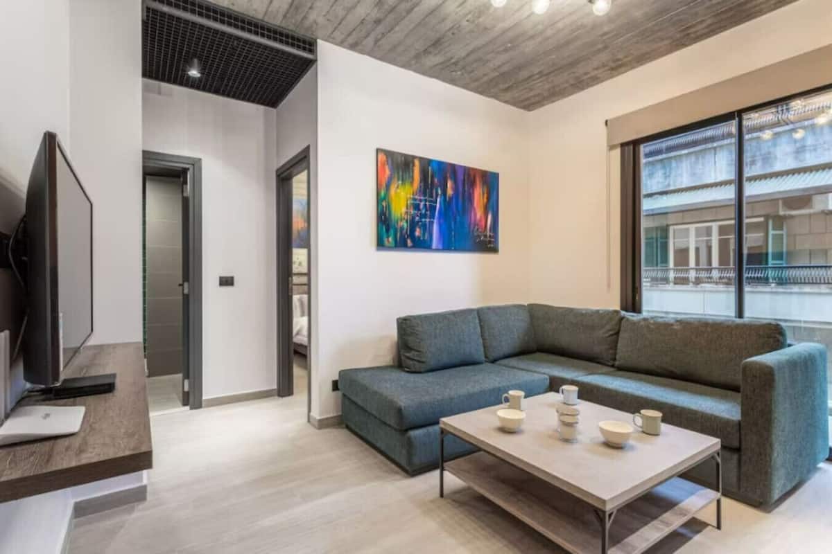 Una sala de estar moderna con un sofá gris, una mesa de centro con tazas, un televisor sobre un mueble de madera y un cuadro abstracto colorido en la pared.