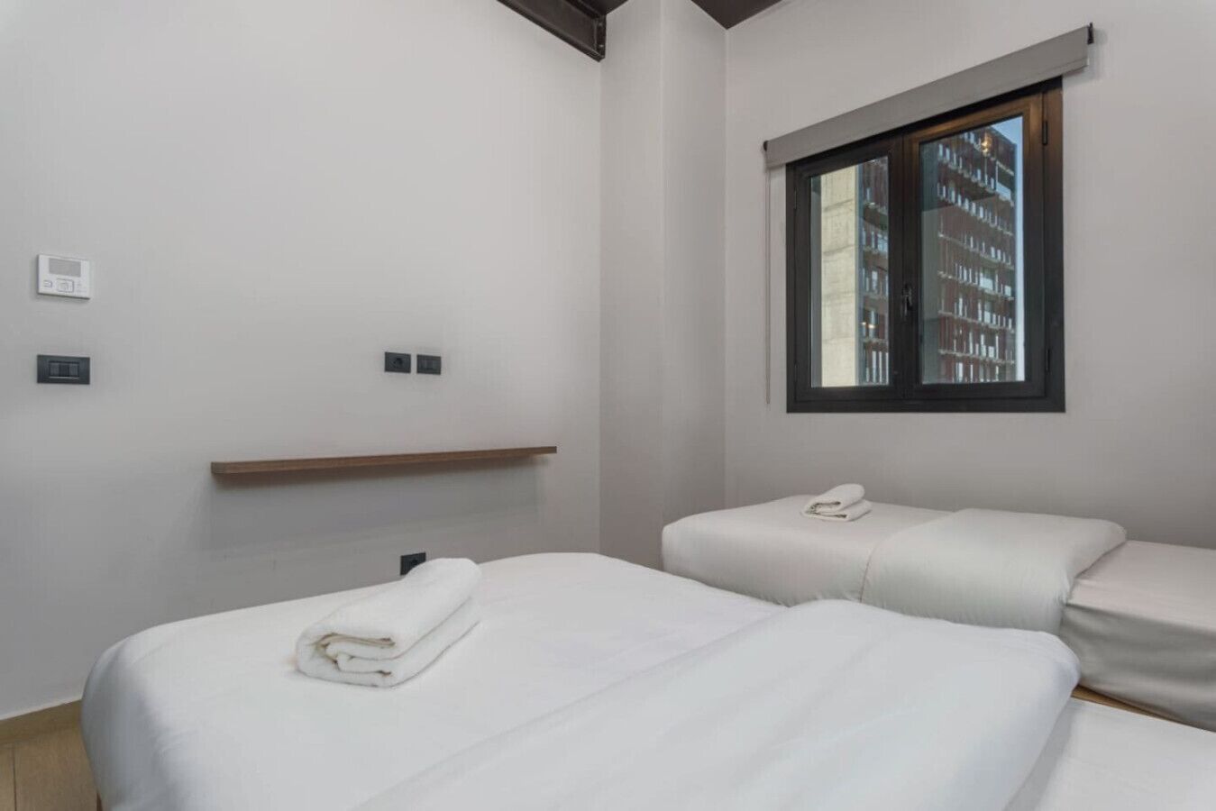 Una camera d'hotel con due letti, una finestra vista edifici e uno scaffale in legno.