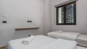 A hotel room with two beds, a window with a view of buildings, and a wooden shelf.
