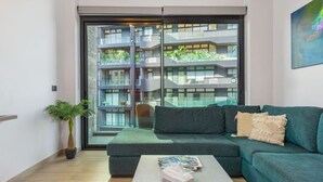 A modern living room with a teal sofa, a coffee table, and a large window offering a view of a multi-story building with balconies. - Amine Apart-Hotel by Stayinn (Beirut)