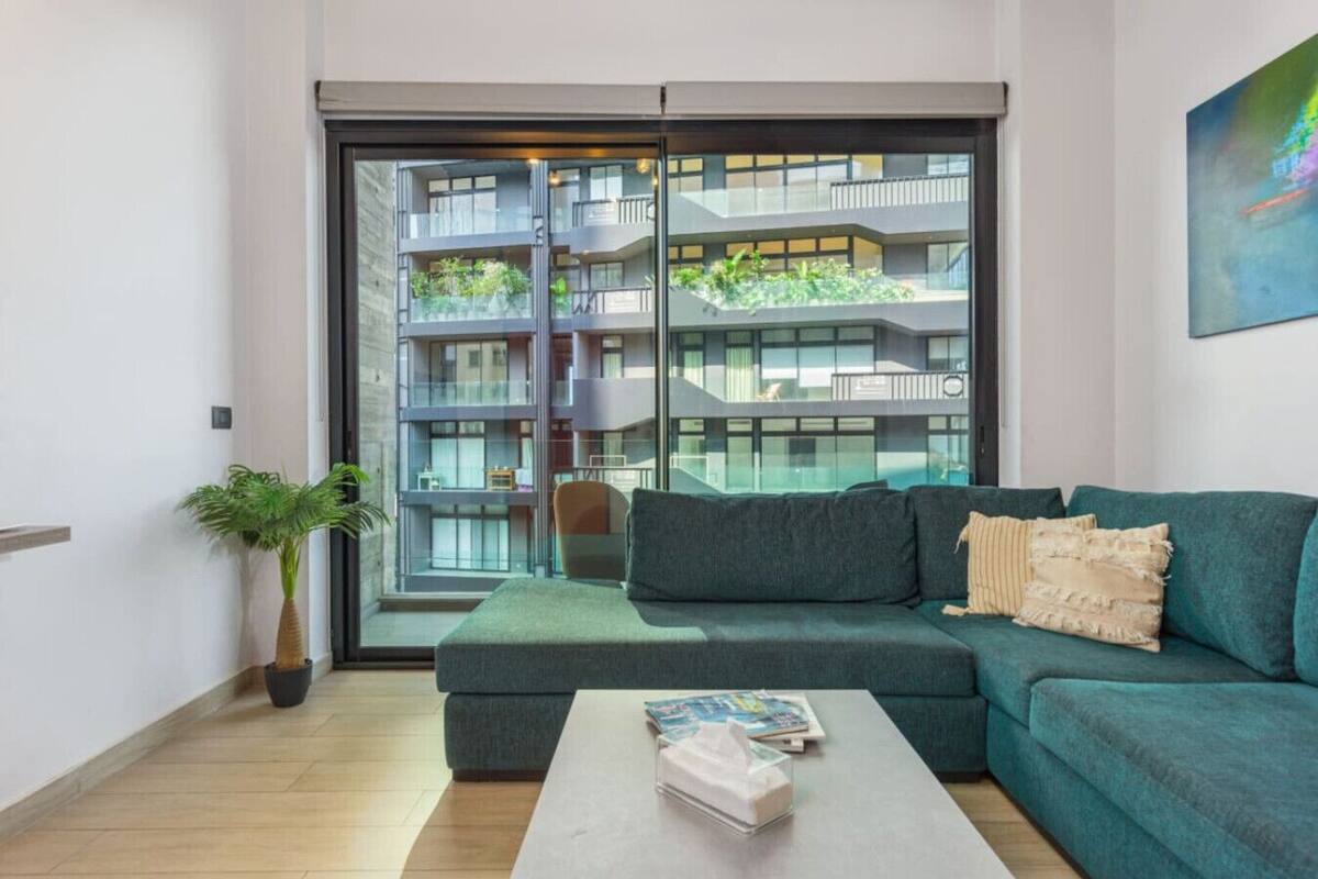 Una sala de estar moderna con un sofá azul turquesa, una mesa de centro y un amplio ventanal que ofrece vistas de un edificio de varios pisos con balcones.