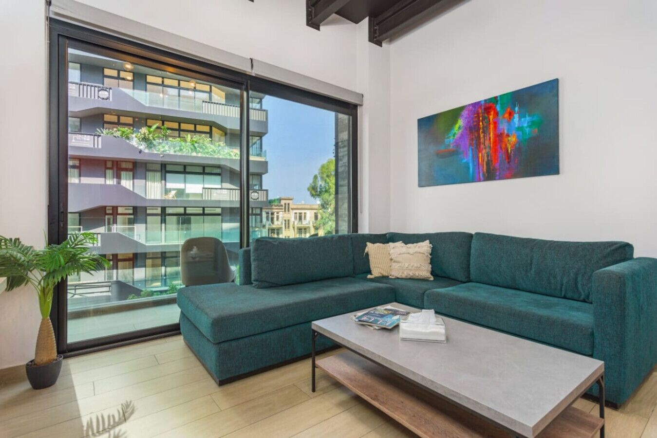 A modern living room with a teal sofa, a coffee table, and a large window offering a view of a building with balconies.