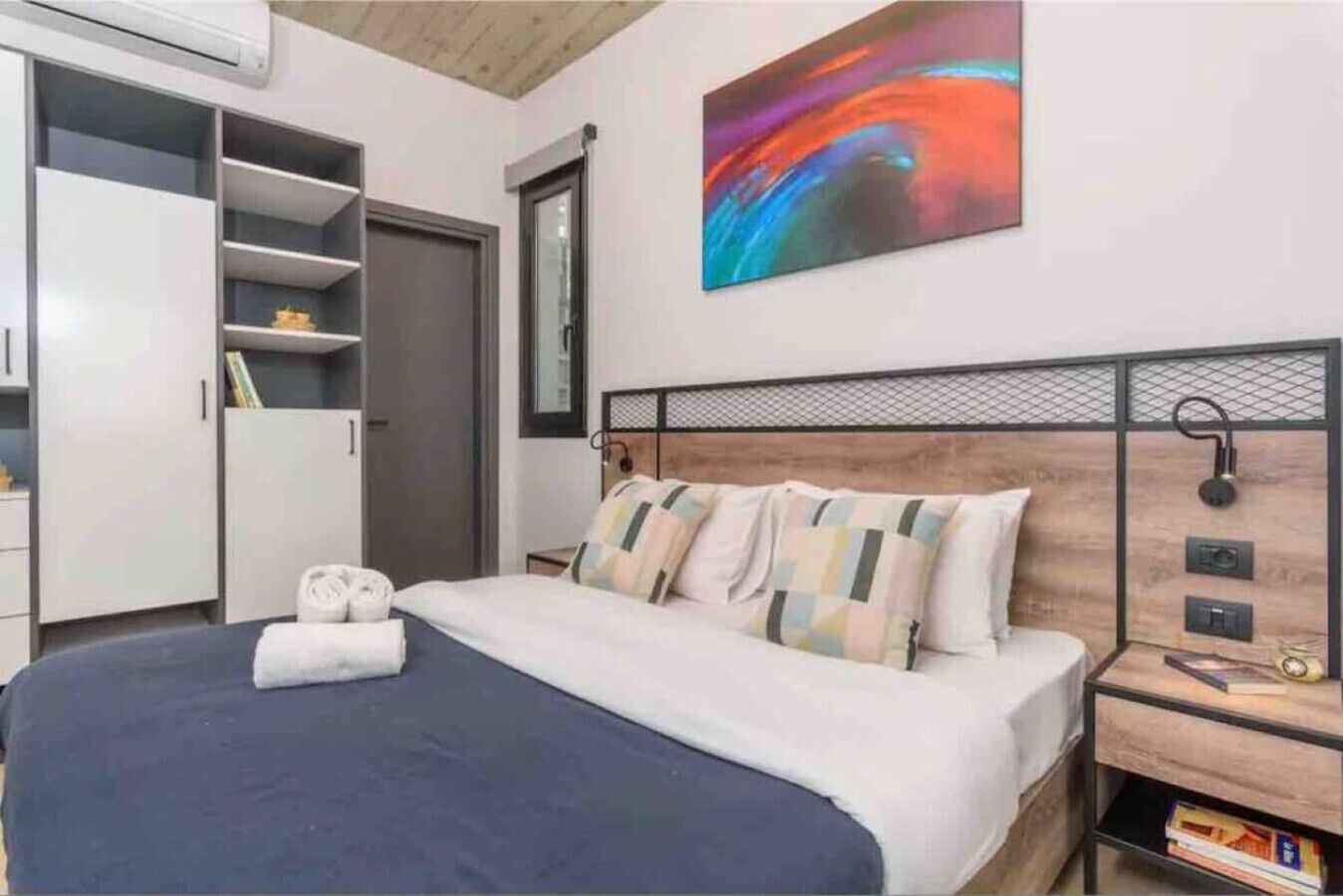 A bedroom with a bed, a wardrobe, a bedside table with books, and a colorful abstract painting on the wall.
