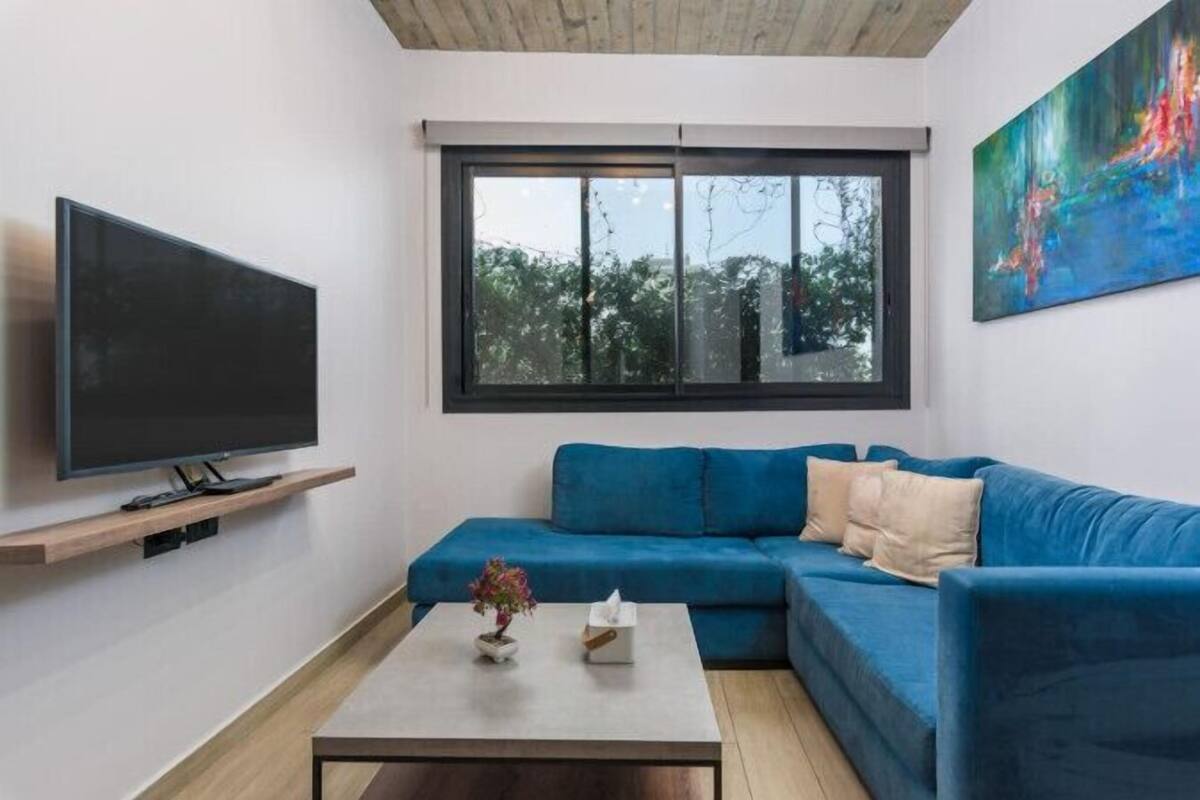 Una sala de estar moderna con un sofá azul, un televisor de pantalla plana, una mesa de centro con un jarrón y una caja de pañuelos, y un amplio ventanal con vistas a árboles.
