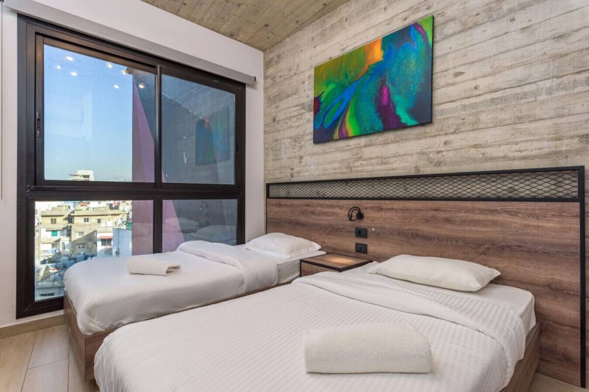 Un dormitorio moderno con dos camas, un ventanal grande y un cuadro abstracto colorido en la pared.