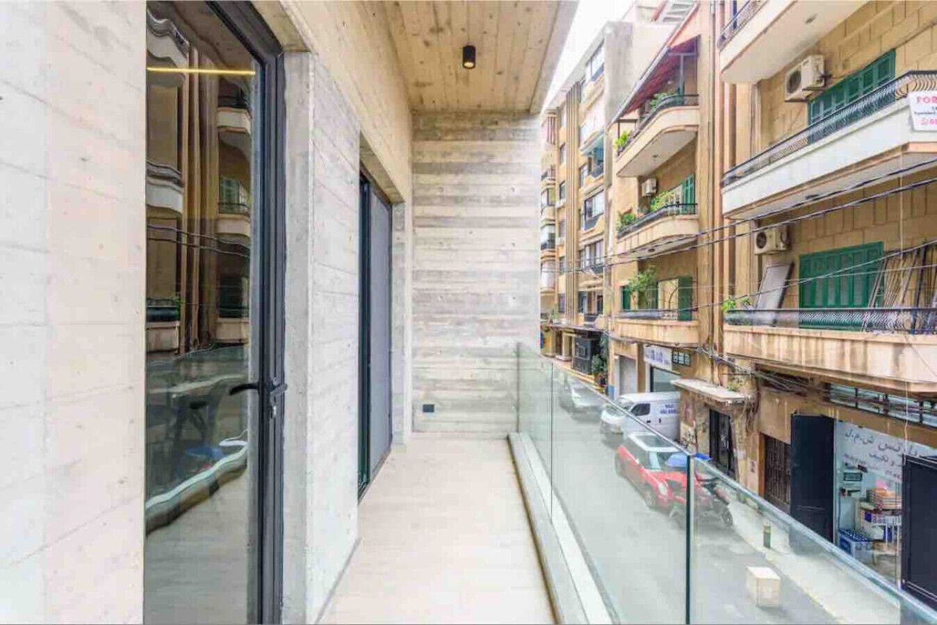 Un pasillo estrecho con barandilla de vidrio, que conduce a un edificio con varios balcones y coches aparcados.