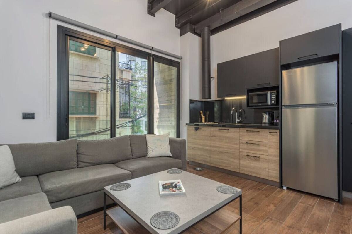 Una sala moderna con un sofá gris, una mesa de centro con revistas, y una zona de cocina con un refrigerador de acero inoxidable, microondas y armarios de madera.