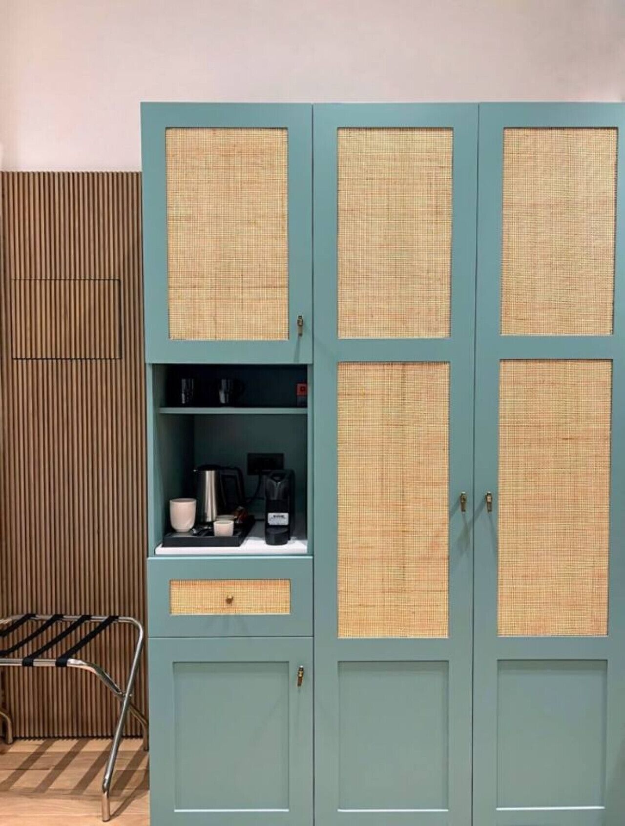 A teal-colored cabinet with woven panels and a shelf containing a coffee maker, cups, and a kettle.