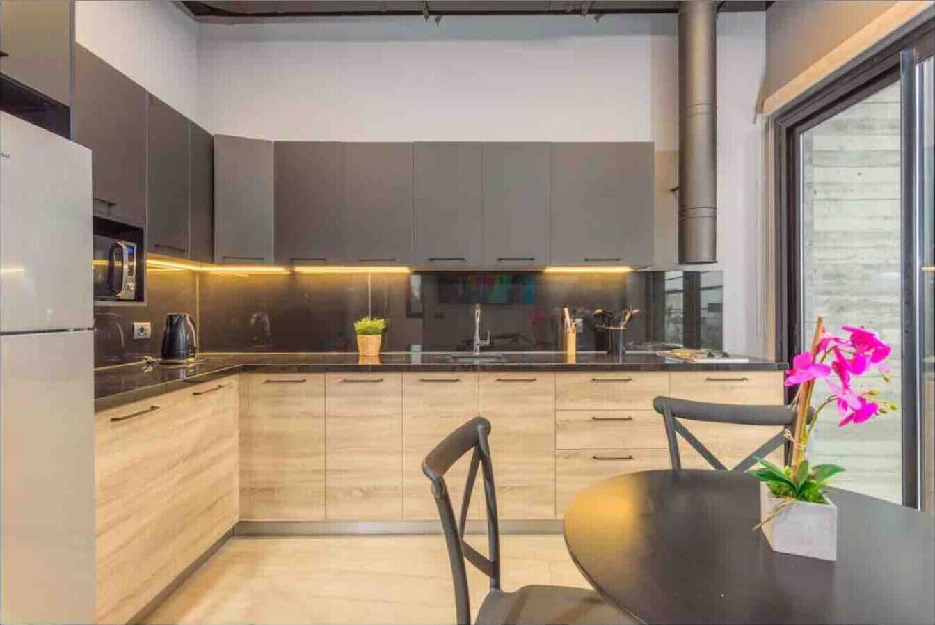 Een moderne keuken met lichte houten kastjes, een zwart aanrecht en een eethoek met een tafel en stoelen.