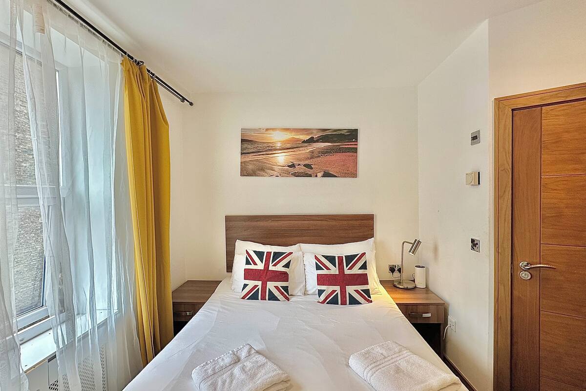 Ein Hotelzimmer mit einem Bett, zwei Kissen mit Union-Jack-Muster, einem Nachttisch mit Lampe und einem Strandbild an der Wand.