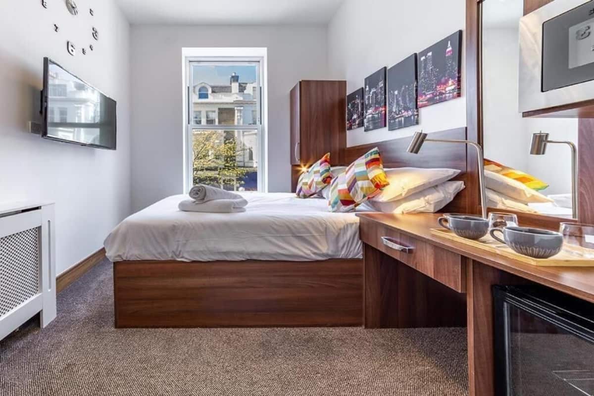 Ein modernes Hotelzimmer mit einem hölzernen Bett, einem flachbildfernseher an der Wand, einem Fenster mit Blick auf die Stadt und einem Schreibtisch mit Lampe und Tasse.