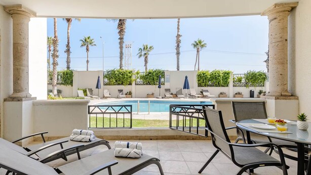 A sunlit outdoor seating area with lounge chairs and a table, overlooking a pool and palm trees.