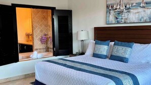 A hotel room with a large bed, a painting of sailboats, a bathroom with a bathtub, and a window with a view of the sea.