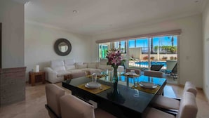 A modern living room with a dining area, a sofa, a coffee table, and a view of a pool and greenery.