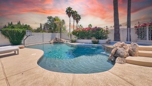 A swimming pool with a basketball hoop, surrounded by palm trees and a sunset sky.